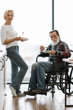 An elderly couple shares a meal, with the man seated in a wheelchair and the woman standing beside him