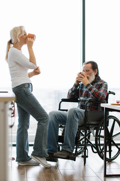 A senior couple shares a quiet moment drinking coffee together by a large window, one seated in a wheelchair