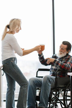 A woman offers a cup of coffee to an elderly man sitting in a wheelchair near a large window