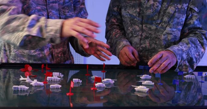 Two uniformed military personnel move small ship models on a glowing digital map table during a naval strategy planning exercise.