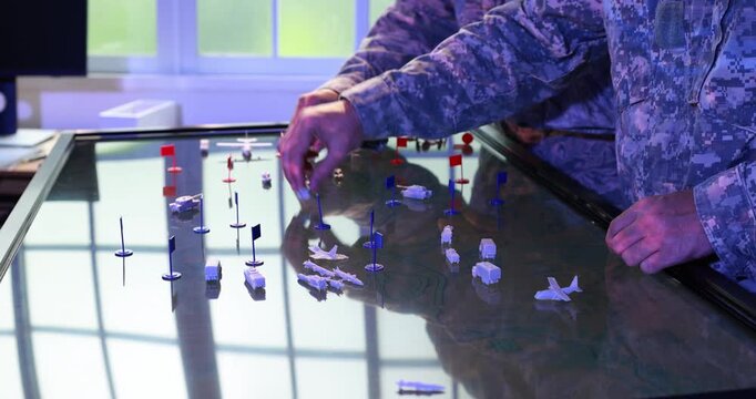 Military officer uses a digital tabletop map with miniature aircraft, ships and markers during a tactical planning exercise in a modern command room.