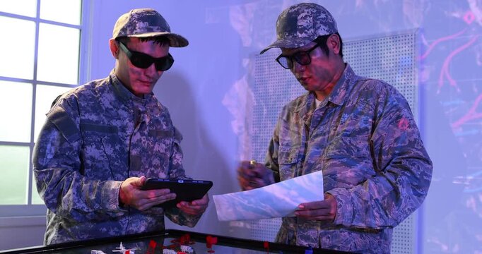 Two soldiers in camouflage uniforms wear protective glasses while reviewing a map and using a handheld device in a military training room.