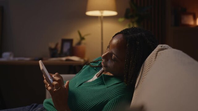 Young adult African American woman lying down, scrolling on smartphone late at night, looking exhausted. Footage represents digital burnout, sleep deprivation, and mental wellbeing concepts