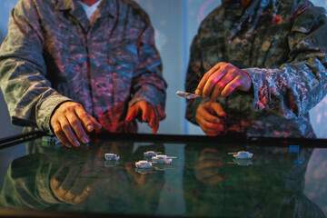 Naklejka premium Two camouflage-clad soldiers use a digital touchscreen map with miniature military vehicles during a tactical planning exercise in low colorful light.