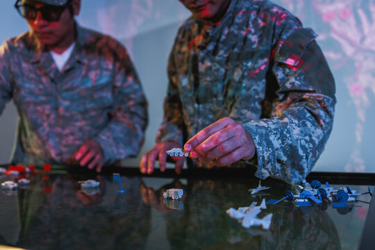 Two uniformed soldiers play a tabletop war game with miniature figures and dice under dramatic red and blue lighting.