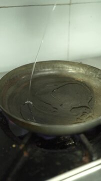 Close-up vertical shot of cooking oil being poured from a stainless steel ladle into a heavy iron skillet over an active gas burner flame in a professional kitchen setting.