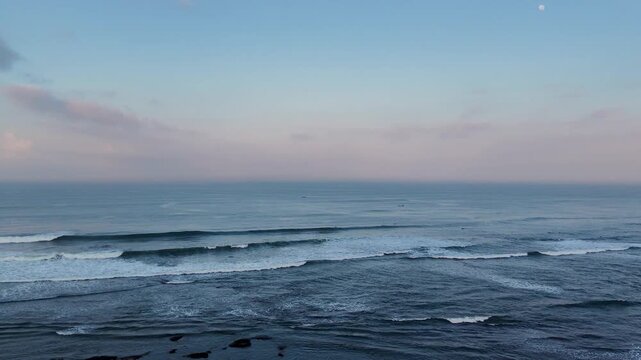 Peaceful ocean view from Kedungu Beach, Tabanan, Bali at dawn. Soft pastel sky with moon, rolling waves breaking over the reef, and dark blue sea