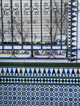 Colorful traditional Spanish ceramic tiles, wrought iron grille and dry plants on a neglected old facade, combining rich decorative patterns with visible signs of decay.