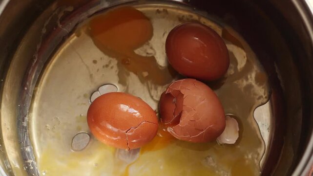 A raw brown chicken egg falls into a metal pot from above alongside several cracked eggs. Upon impact, the shell breaks, causing fresh egg white and yolk to spread across the bottom of the pan.