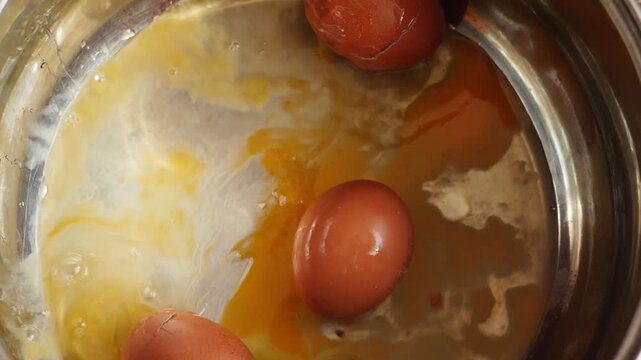A raw brown chicken egg falls into a metal pot from above onto several broken eggs. Upon impact, the shell breaks, causing fresh egg white and yolk to spread across the pan.