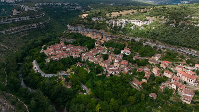 peaceful aerial view of a small village nestled within forested hills in rural, mountainous terrain with red-tiled roofs and stone buildings, Risco los camellos, Cascada de Orbaneja del Castillo