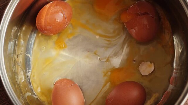 A raw brown chicken egg falls into a metal pot from above onto several broken eggs. Upon impact, the shell breaks, causing fresh egg white and yolk to spread across the pan.