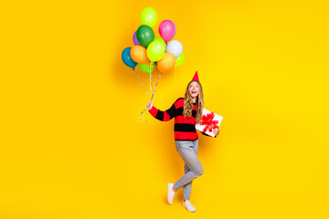 Cheerful young woman holding colorful balloons and a gift while wearing a festive party hat with a...