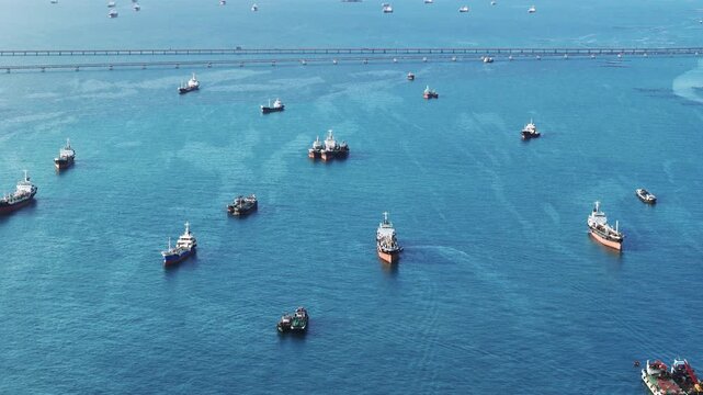 Fleet of oil tanker ships and commercial cargo vessels anchored in the deep blue sea near a long bridge. Global maritime logistics, international shipping trade, and energy transport.
