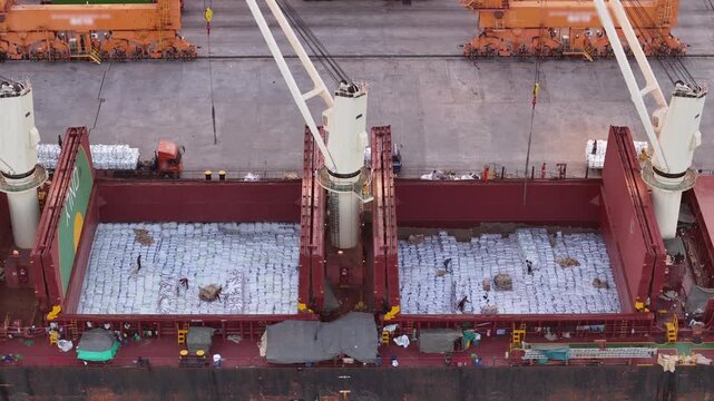 Cargo ship using deck cranes to load white bags from trucks at the port terminal. Maritime logistics, industrial shipping, and international freight transport operations.