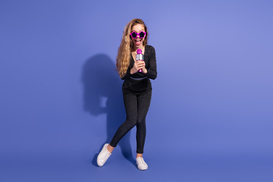 Young woman with pink glasses singing into microphone in striped jumpsuit in a playful blue studio portrait