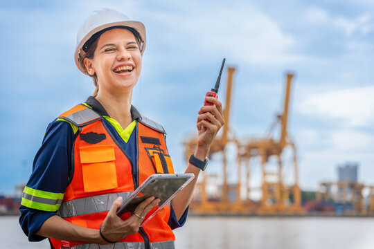 Smiling female port engineer holding walkie talkie and digital tablet at container terminal, Happy woman logistic manager with radio device, Confident industrial worker in safety vest at shipping port