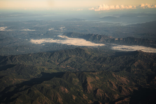 Aerial view of the rugged mountainous landscape of Madagascar, featuring deep valleys, rolling hills, and low-hanging clouds under a soft golden light, Madagascar.