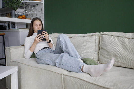 Relaxing woman using digital reader electronic book at home. Beautiful model with long hair in white and green interior indoor