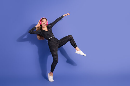Young woman in striped jumpsuit wears pink headphones jumping and smiling against a violet background for a stylish fun lifestyle and fashion advertising image