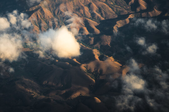 Aerial view of the rugged mountainous landscape of Madagascar, featuring rolling hills illuminated by golden hour light with scattered clouds casting shadows over the terrain, Madagascar.