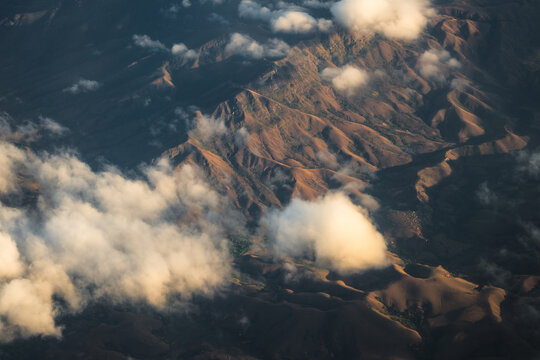 Aerial view of rugged mountainous terrain illuminated by golden hour light with scattered clouds casting shadows over the landscape, Madagascar.