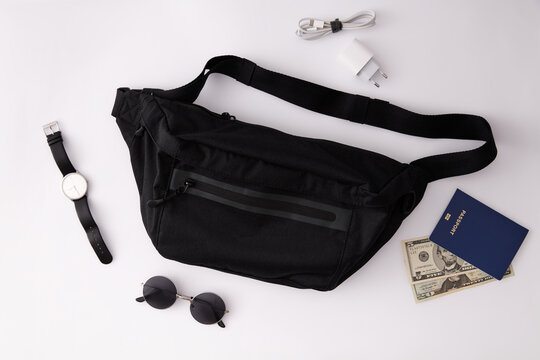 Black waist bag with travel accessories, passport, money, sunglasses, and charger on white background