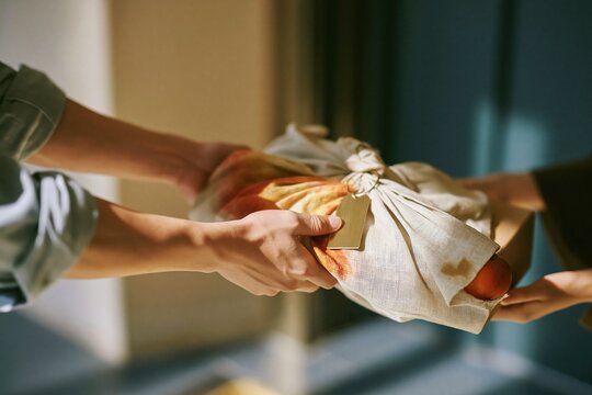 Close up of hands exchanging a fabric wrapped gift with a persimmon, featuring textured ramie cloth in a modern lobby for gifting and seasonal campaigns with copy space