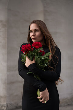 Brunette female holding crimson flower bundle while standing gracefully against minimalist textured architectural concrete background