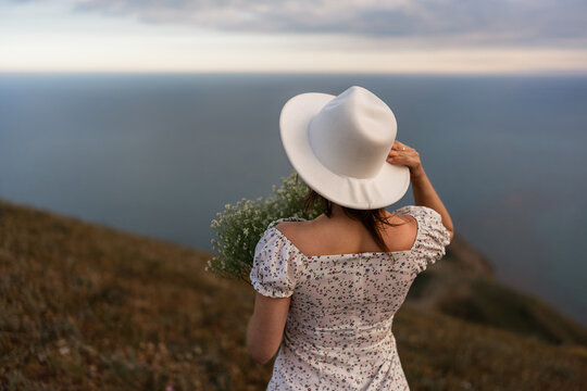 Young woman holding baby breath flowers wears wide brimmed hat while standing on a high grassy coastal cliff during sunset