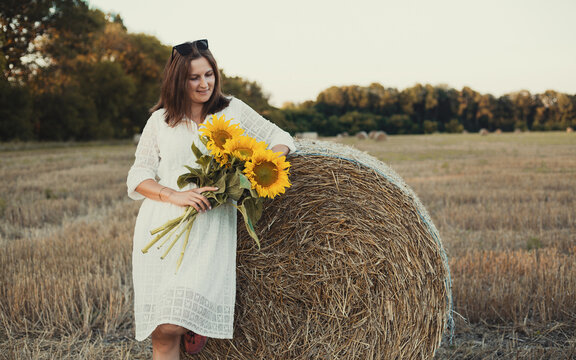 woman in a white dress holding a bouquet of wild flowers with sunflowers in a mowed field with straw bales