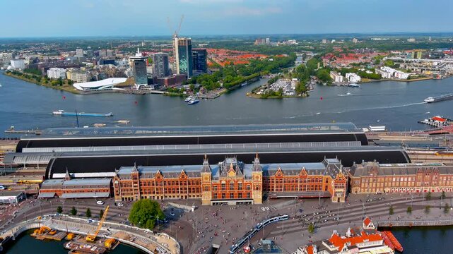Aerial drone view of Amsterdam Centraal railway station with trams and travelers on Stationsplein, platform roofs, ferries on the IJ river and the Amsterdam Noord skyline, Netherlands.