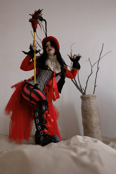 Circus ringmaster in red top hat and striped corset kneeling on sand with feathered staff