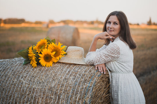 woman in a white dress holding a bouquet of wild flowers with sunflowers in a mowed field with straw bales