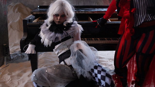 Harlequin performer sitting on sand at the foot of a piano with red gloved hand above