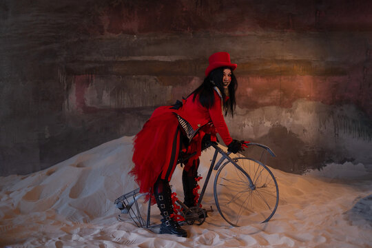 Woman in red circus costume with vintage bicycle on sand in derelict hall