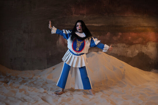 Performer in blue and white pierrot dress with arms outstretched on sand dune