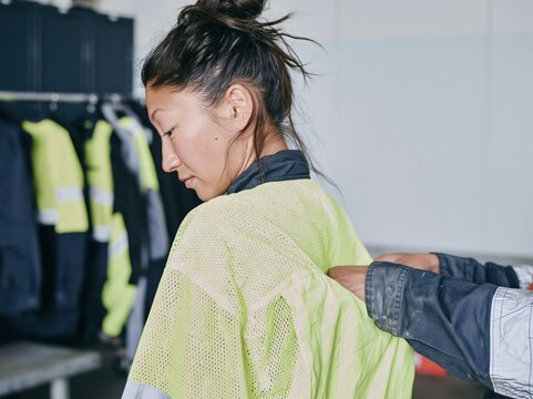 East Asian woman wearing a fluorescent hi vis vest being adjusted by a mentor in a gear room, for workwear and women in trades campaigns