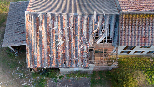 Aerial view of a rural house roof with damaged terracotta tiles and exposed structural beams caused by a storm, with debris on the ground in Castelvetro Piacentino, Emilia-Romagna, Italy.