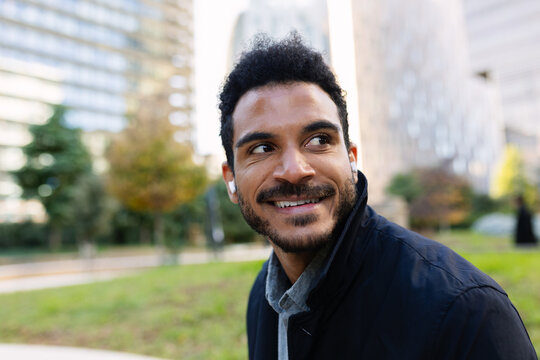 Portrait of young adult Latin American businessman wearing earbuds outdoors. Relaxed professional enjoying audio while surrounded by greenery. Business and people concept.