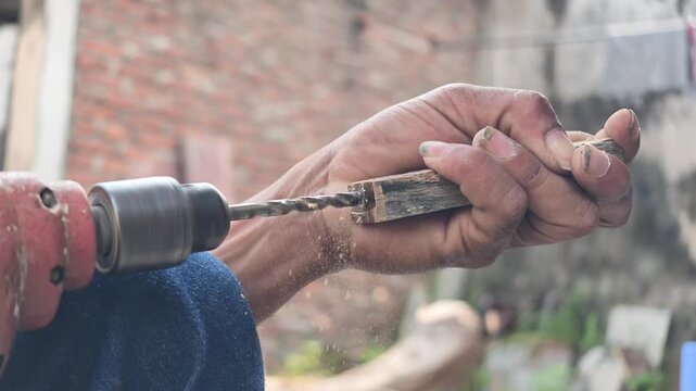 Close-up shot of artisan hands operating an electric power drill to bore a deep hole into a small square timber block for a custom tool handle.
