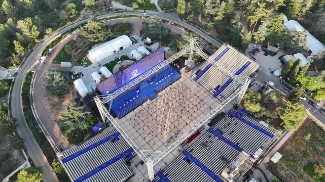 High angle aerial view of an open air event venue stage and seating structure surrounded by green forest trees during sunny daylight in Jerusalem