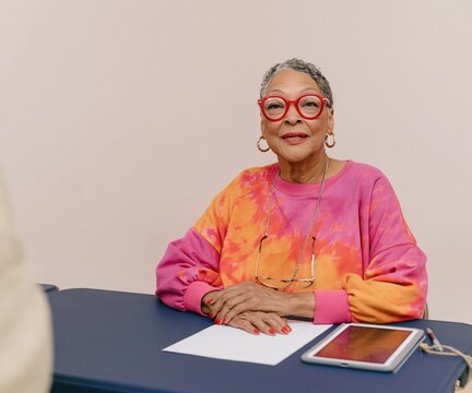 Senior Black woman poll worker sitting at a check-in table with a blank paper roster and tablet, featuring sharp textures for election communication and recruitment
