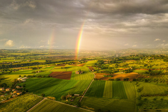 Aerial view of a vibrant rainbow arching over the lush green agricultural fields and rolling hills of the countryside in Corridonia, Marche, Italy.