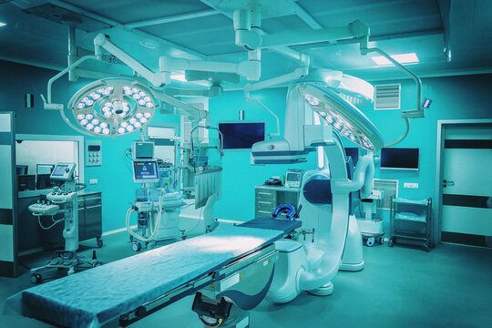 Surgical lamps hang over the table in a hybrid operating room of a new hospital. The image is toned in cold cyan with visible grain, noise and a vignette filter. Cinematic medical mood