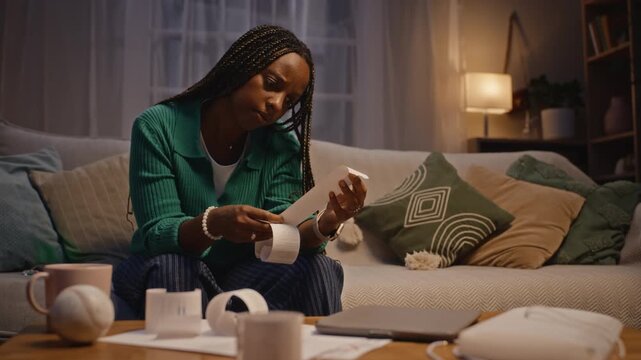 African American woman sitting on sofa at night, examining paper receipts and showing exhaustion. Video is suitable for financial planning services, debt relief ads, or mental health campaigns
