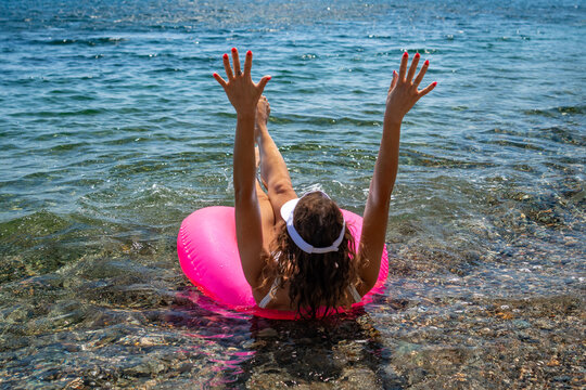 Floating joyfully on a bright pink tube in the sparkling sea while raising arms during summer vacation