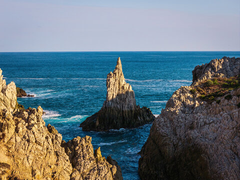 Aerial view of the Dedo de Dios rock formation rising from the deep blue Pacific Ocean, surrounded by rugged coastal cliffs and golden sunlight in Maruata, Michoacan, Mexico.