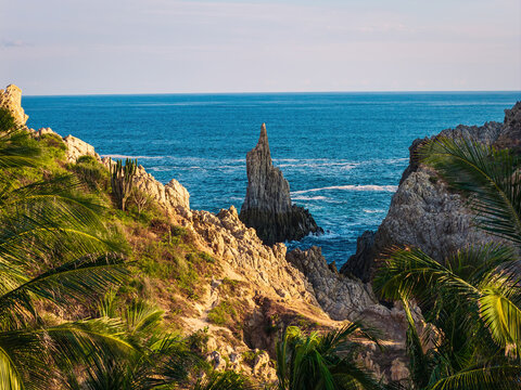 Aerial view of the Dedo de Dios rock formation rising from the Pacific Ocean, surrounded by rugged cliffs, palm trees, and coastal vegetation in Maruata, Michoacan, Mexico.