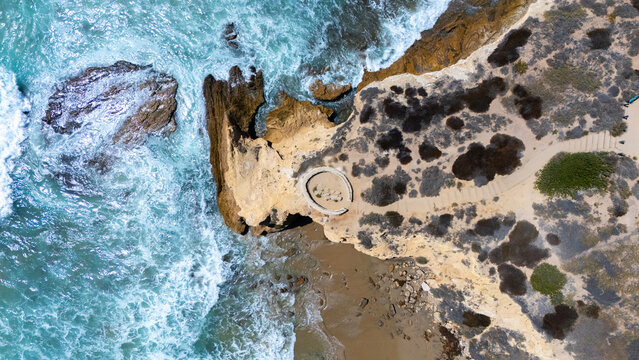 Aerial view of the rugged coastline at Crystal Cove, featuring dramatic cliffs, crashing turquoise waves, and a circular stone structure in Newport Beach, California, United States.
